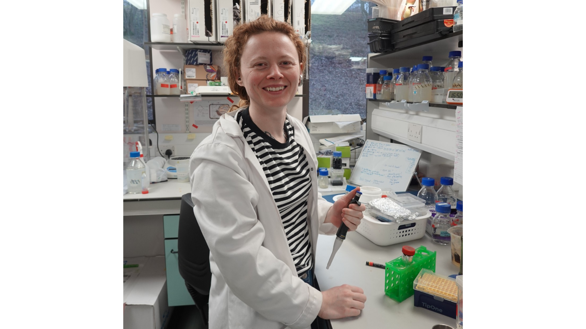 Alice Fidell, a Technician at the Department of Biosciences, Durham University, sits at a laboratory bench, smiling as she holds a pipette above a set of small sample tubes and lab equipment. Wearing a lab coat and surrounded by bottles, racks, and notes from ongoing experiments, she works with the tools used to study how plants respond to copper toxicity at the molecular and developmental level.