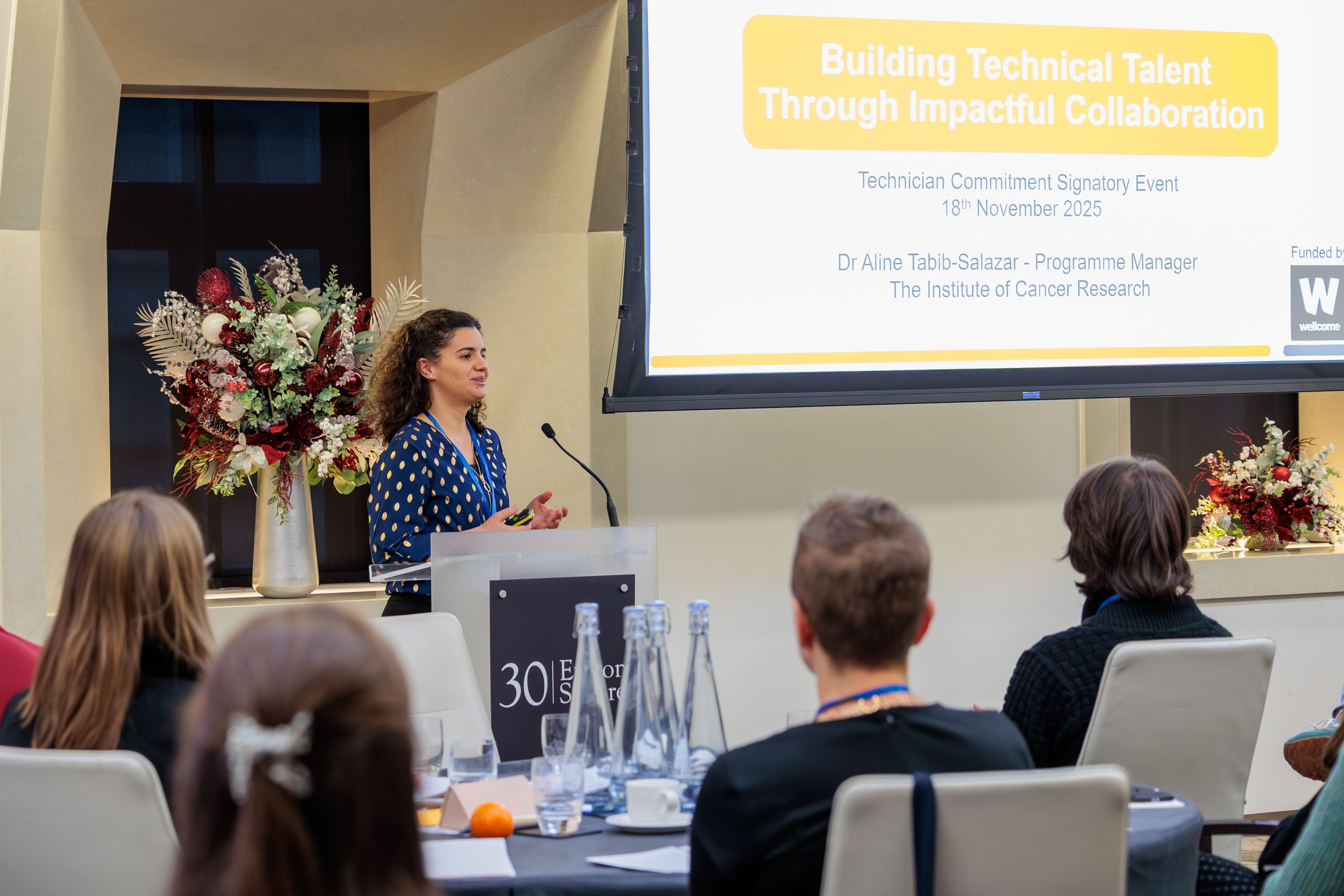 Dr Aline Tabib-Salazar, Technician Career Pathways Programme Manager at The Institute of Cancer Research standing at a podium with a microphone, in front of a PowerPoint slide which reads: 'Building Technical Talent Through Impactful Collaboration . Technician Commitment Signatory Event. 18th November 2025.  Dr Aline Tabib-Salazar, Programme Manager. The Institute of Cancer Research'.  Audience members can be seen sitting at roundtables facing Aline.