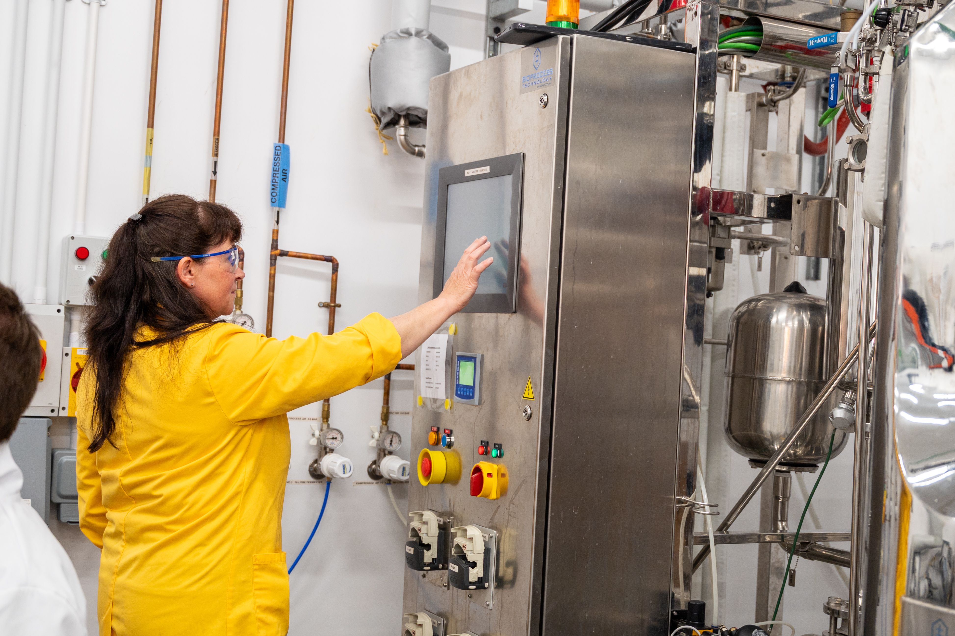 Sian Jones, a Senior Research Technician at IBERS, stands in a laboratory beside large-scale research equipment, operating a touchscreen control panel while monitoring the system. Wearing protective glasses and a lab coat, she manages specialised processing equipment used in bioresource and biorefining research.