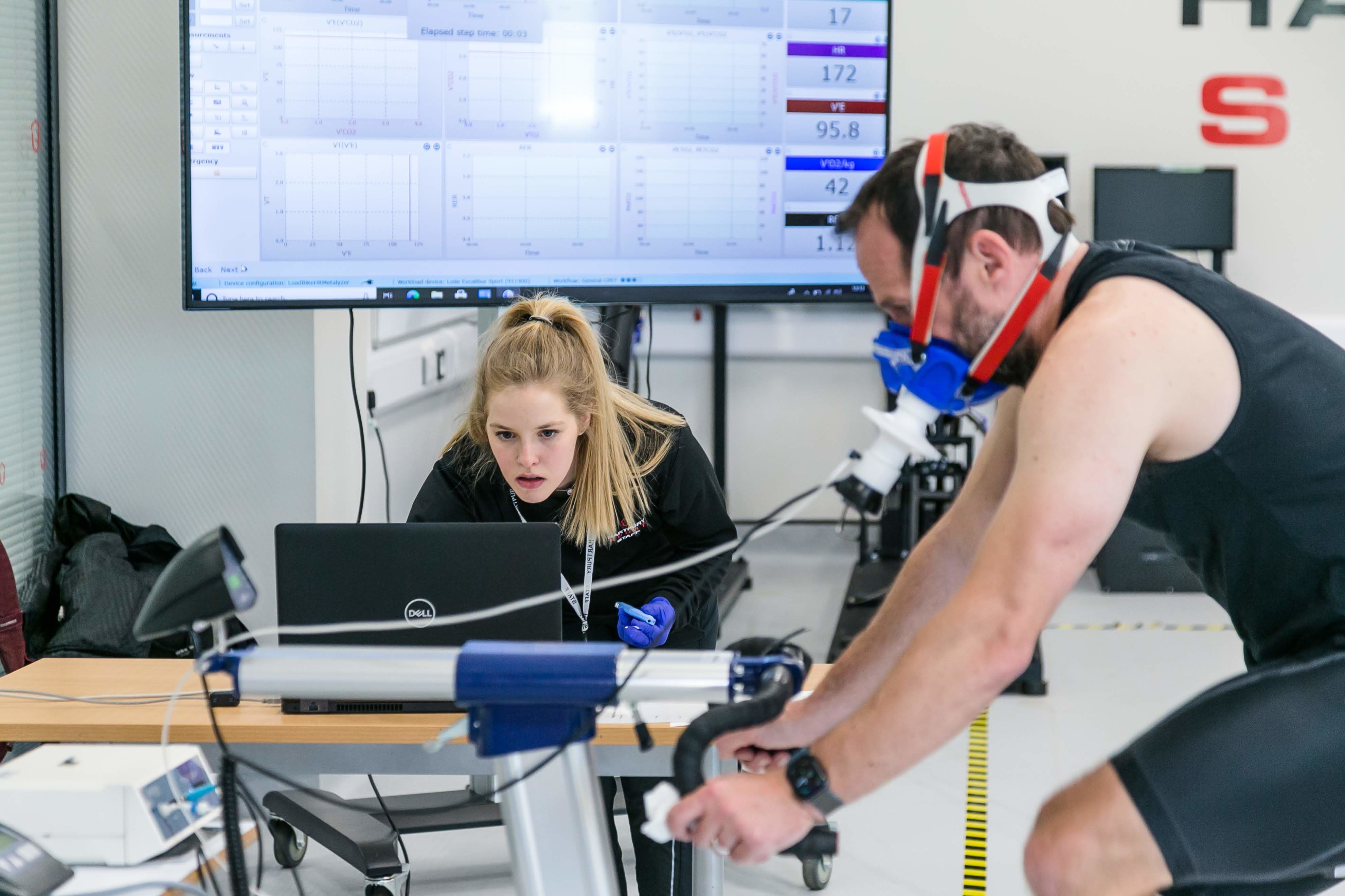 Amelia Dingley, a Technical Manager at Brunel University, London, monitors a cycling lactate test in a sports science laboratory, analysing data on a laptop as a participant pedals on a stationary bike while wearing a respiratory mask. With real-time performance metrics displayed on a large screen behind them, she supports physiological testing that helps researchers and athletes understand endurance, performance, and metabolic responses to exercise.