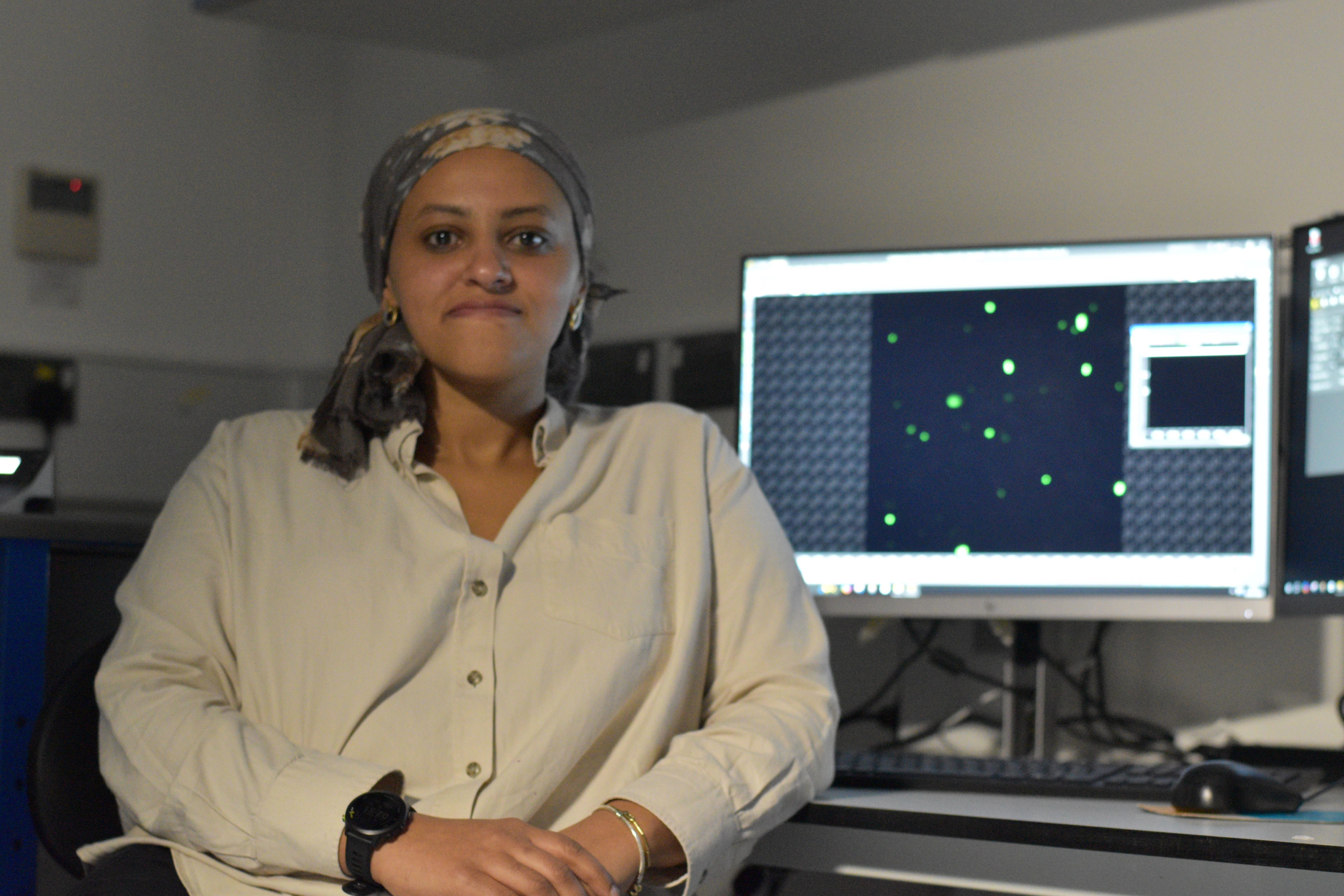 Mezida Saeed, Advanced Imaigng Specialist at COMPARE Advanced Imaging Facility, University of Birmingham, sits at a workstation in an imaging laboratory, with a monitor behind her displaying fluorescent microscopy images of cells. Working with advanced imaging software and equipment, she analyses detailed biological data to support cutting-edge research.