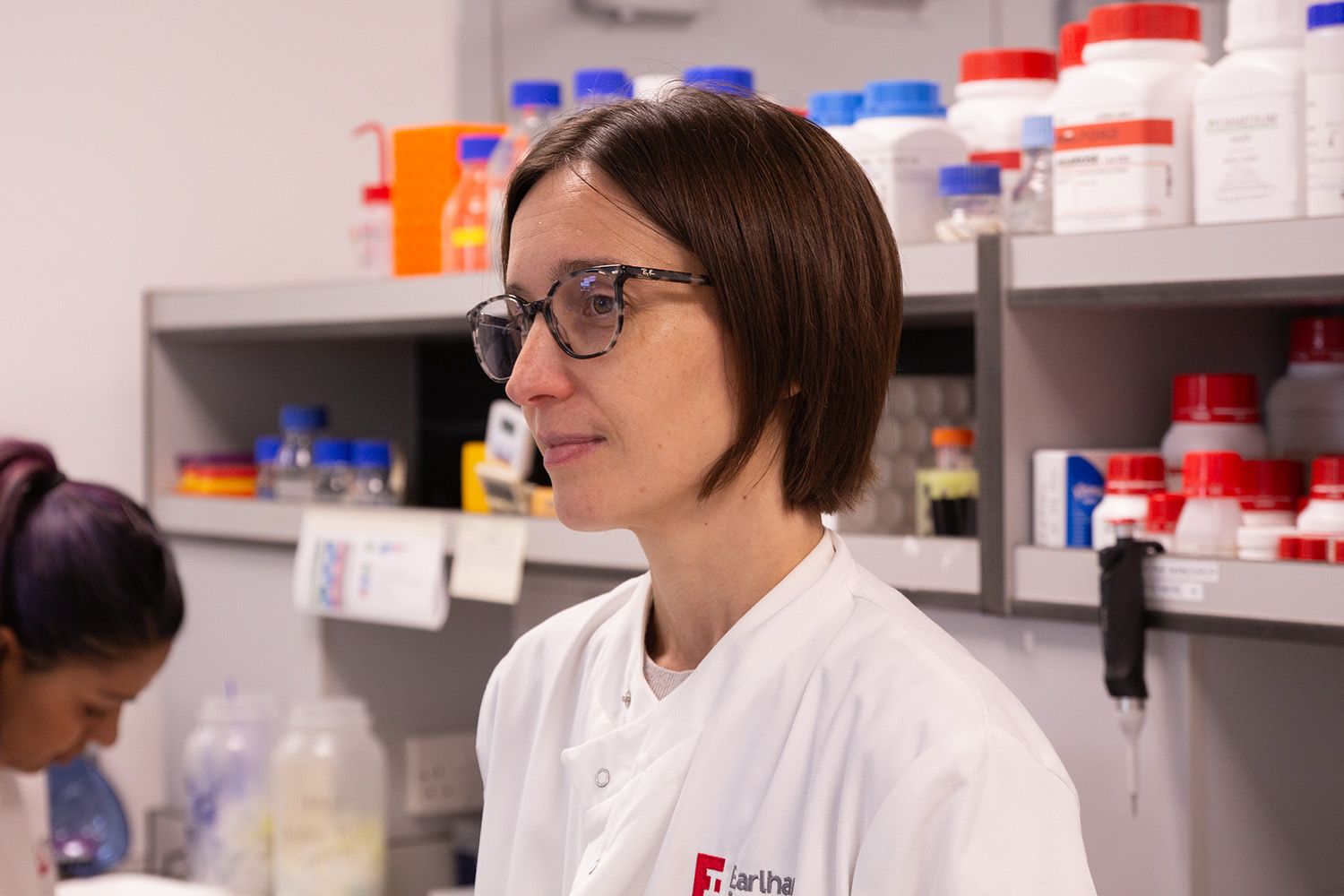 Dr Carolina Grandellis, Head of the Earlham Biofoundry, Earlham Institute, stands in a laboratory wearing a white lab coat and glasses, focused as she works among shelves of reagents and laboratory supplies. Her work at the biofoundry supports advanced biological research, helping scientists design, build, and test biological systems using automated and high-throughput technologies.