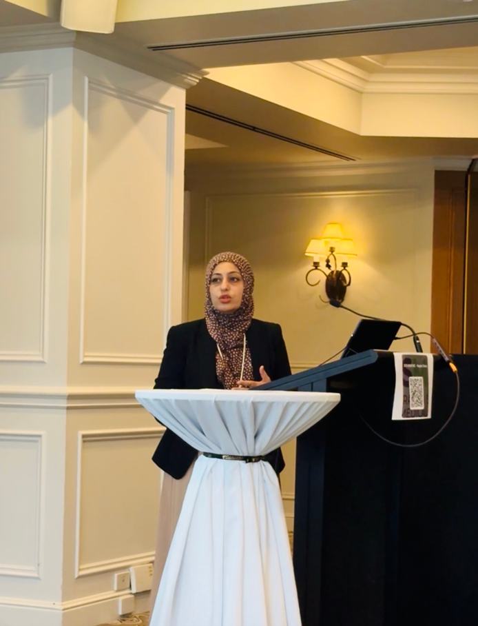 Farah Zahir, Head of Statistics and Principal Data Scientist at QCIF, stands at a podium delivering a presentation, speaking to an audience while gesturing thoughtfully. Dressed in professional attire, she presents statistical insights and data-driven research, reflecting her leadership role in biostatistics and data science.