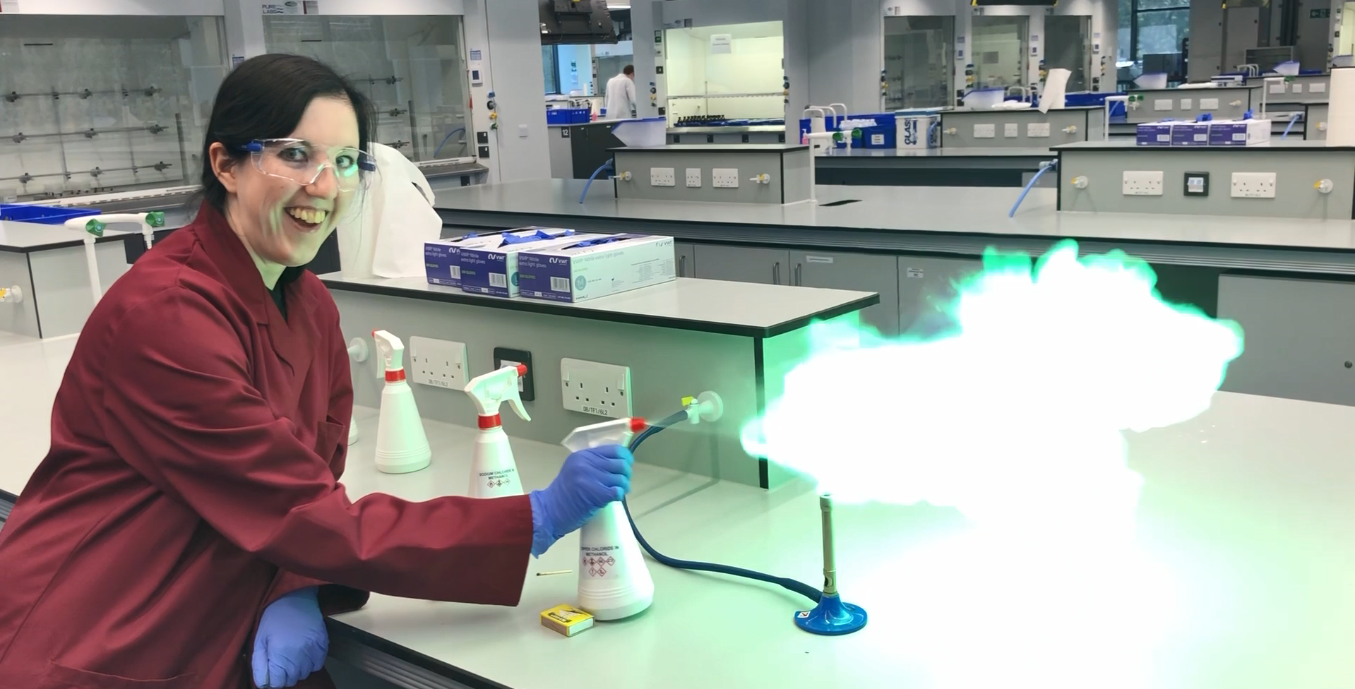 Jasmine Downes, Senior Laboratory Technician at University of East Anglia,  demonstrates a flame test in a teaching laboratory, carefully using a spray bottle to produce a bright green flame from a Bunsen burner. Wearing safety glasses, gloves, and a lab coat, she supports practical chemistry teaching by preparing experiments and guiding students through hands-on laboratory techniques.