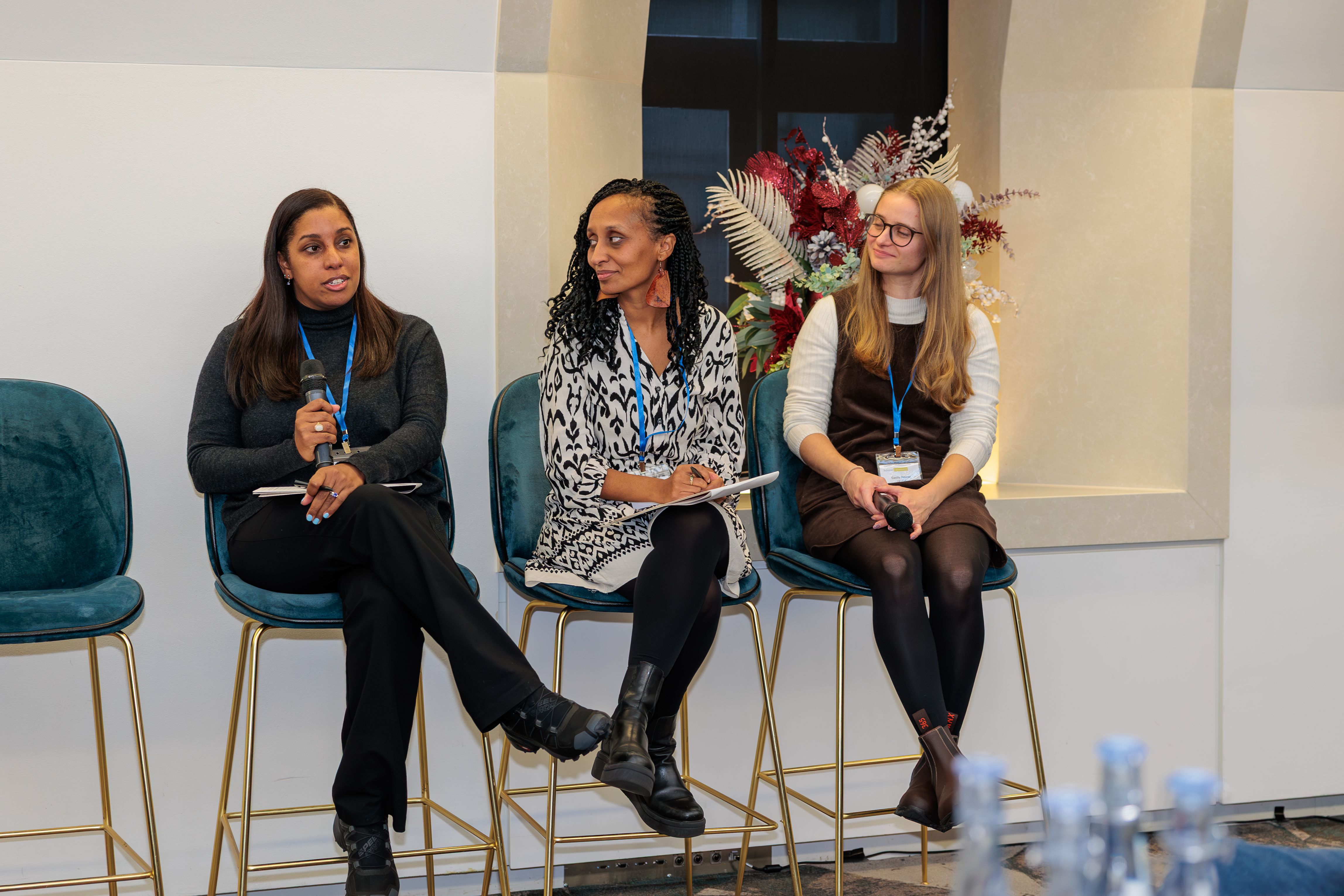 The three speakers who spoke on the international panel at the November 2025 Technician Commitment signatory event (left to right: Brigitte Denis of Malawi Liverpool Wellcome Research Programme; Yara Reis from Global BioImaging, and Cecily Pepper, Research Fellow at UK ITSS) sitting on stools  at the front of the room. Brigitte is speaking into a microphone and responding to a question from an audience member, while the other two panellists have their bodies angled towards her listening to her answer.