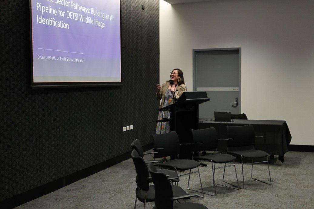 Dr Jenna Wraith, Head of Sustainable Futures Department and Principal Data Scientist at QCIf, stands at a lectern presenting to an audience, gesturing toward a projected slide while holding a microphone. The screen behind her displays a presentation on artificial intelligence and wildlife image identification, reflecting her work applying data science to environmental and sustainability challenges.