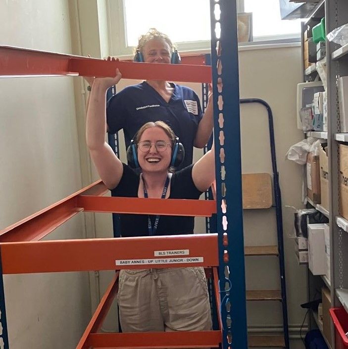 Katy Gough and Jessica Johnston, Clinical Skills Technicians and Trainers from the School of Nursing at the University of Chester, stand in a storeroom between shelving units, smiling as they assemble a large training frame used for clinical skills practice. Wearing protective ear defenders, they work together to organise and prepare equipment that supports hands-on learning for nursing students..