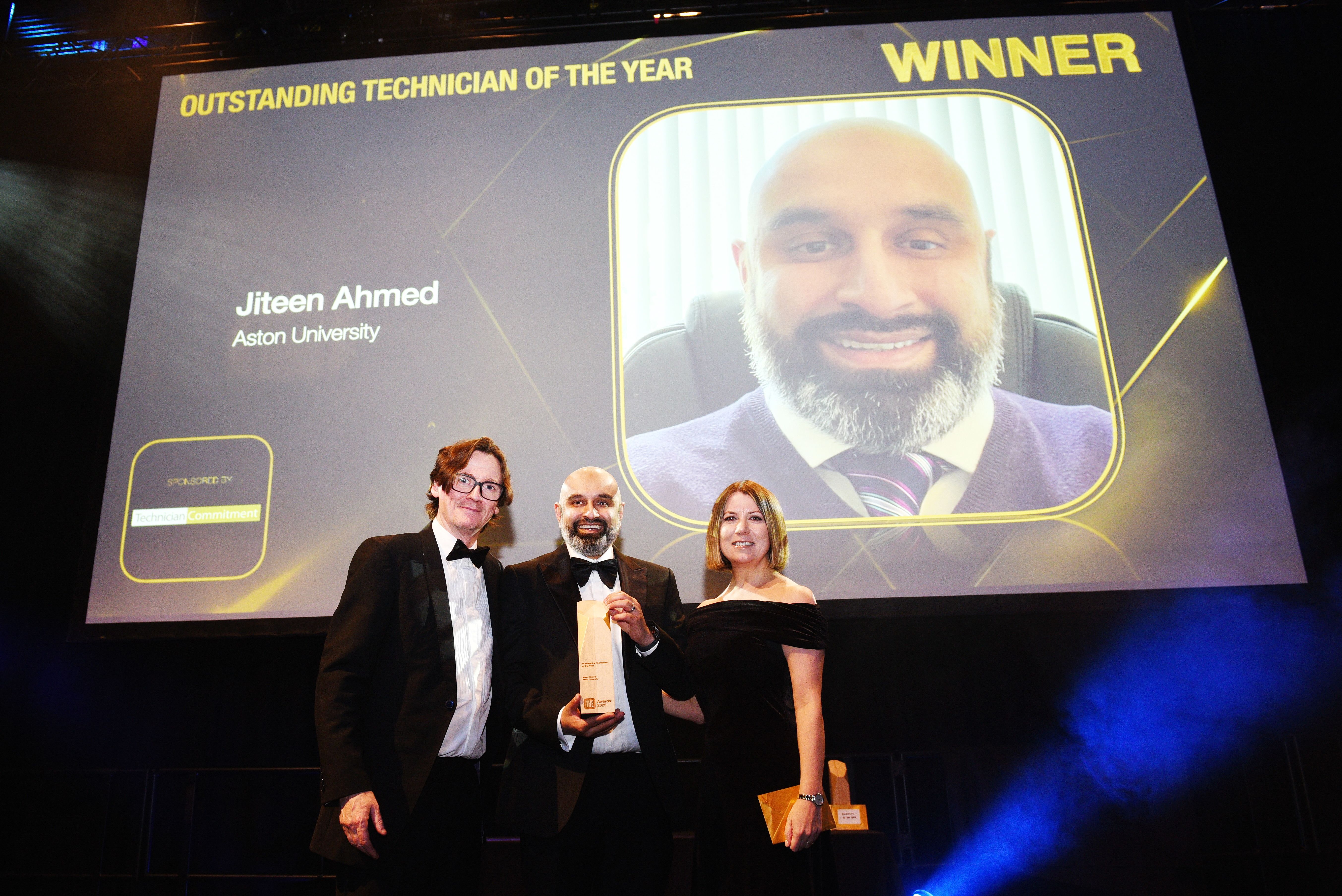 Jiteen Ahmed, Head of Technical Services in the College of Health and Life Sciences at Aston University, stands centre-stage at the Times Higher Education Awards ceremony, holding his Outstanding Technician of the Year Award trophy, flanked by Technician Commitment Programme Lead Kelly Vere MBE and one of the THE Award judges. s. All three are dressed in formal evening wear. Behind them, a large screen displays a photograph of Jiteen alongside bold text reading “Outstanding Technician of the Year – Winner: Jiteen Ahmed, Aston University"