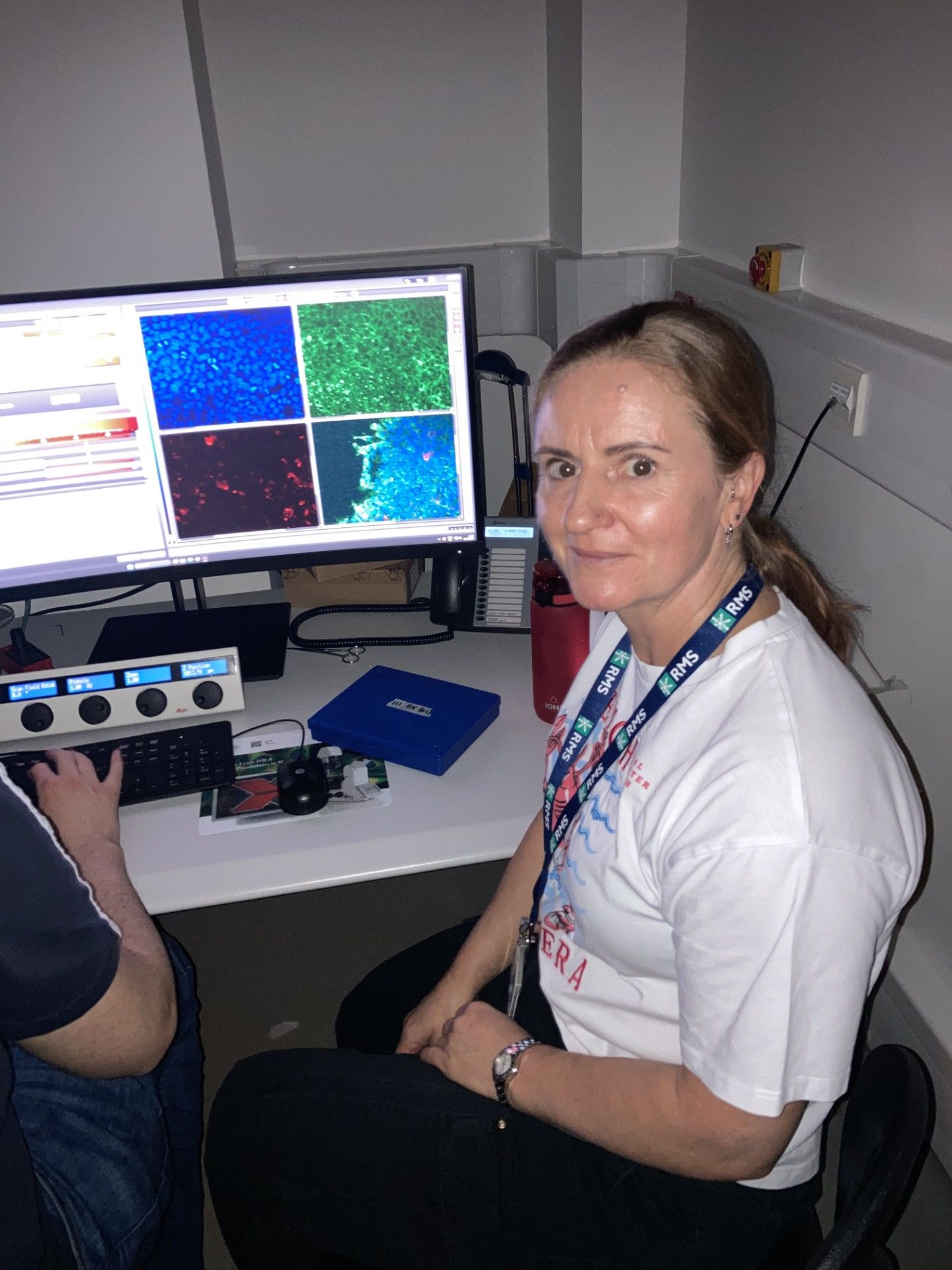 Laura Stirrat, Research Technician/Lead Facility Manager at University of Strathclyde, sits at a workstation beside a computer displaying brightly coloured microscopy images of cells. Wearing a staff lanyard, she works closely with imaging software and specialised equipment used to analyse biological samples and support laboratory research.