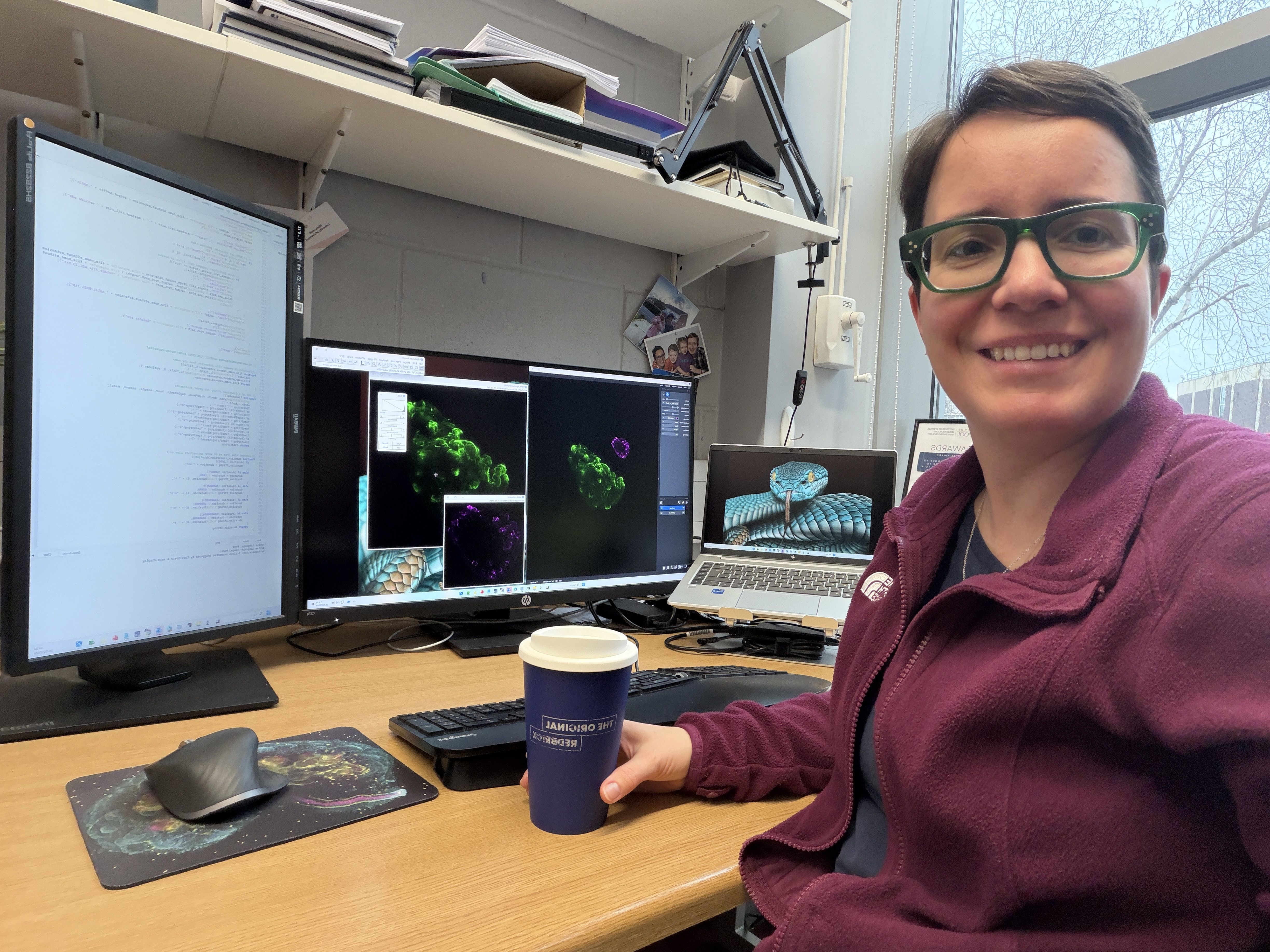 Marie Held, an Image Analyst at University of Liverpool, sits at her desk in a research lab, wearing green-framed glasses and a maroon fleece while holding a takeaway coffee cup. Behind her, several monitors display bright microscopy-style images and analysis software, reflecting her work analysing scientific imaging data.