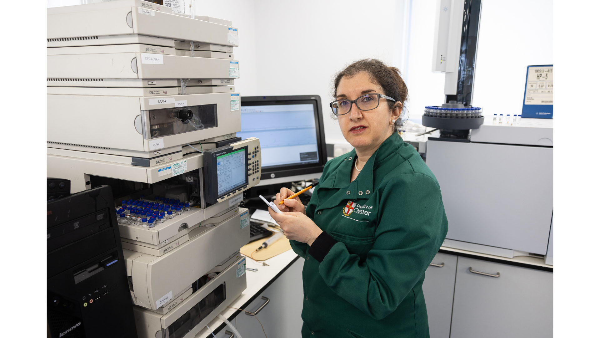 Marlene Barrera, Technical Officer at the University of Chester, stands beside an analytical laboratory instrument, holding a sample vial and making notes as she monitors the system. Wearing a lab coat and surrounded by chromatography equipment and sample trays, she carries out precise analytical work to support scientific research.