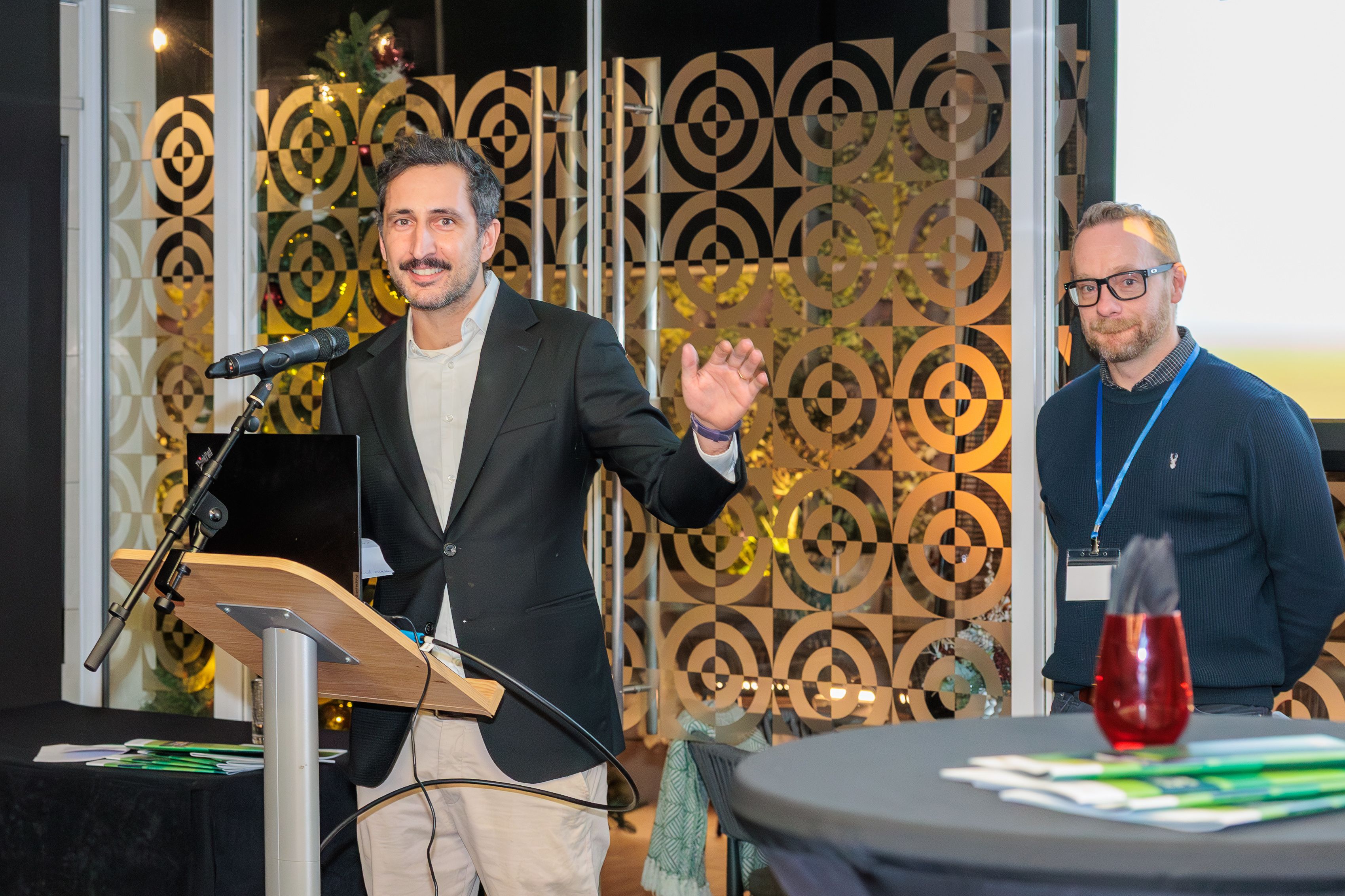 Martin Farley, Associate Director of Environmental Sustainability Programmes at UKRI standing at a podium with a microphone, gesticulating as he speaks. To his left stands Lee Hibbett, Technical Manager at the University of Nottingham who spoke after Martin and whose work on solvent recycling is featured as a case study in the newly launched UK ITSS report "Driving Environmental Sustainability: The Contribution of Technical Professionals in Higher Education and Research". In front of them are hard copies of the report on tables.