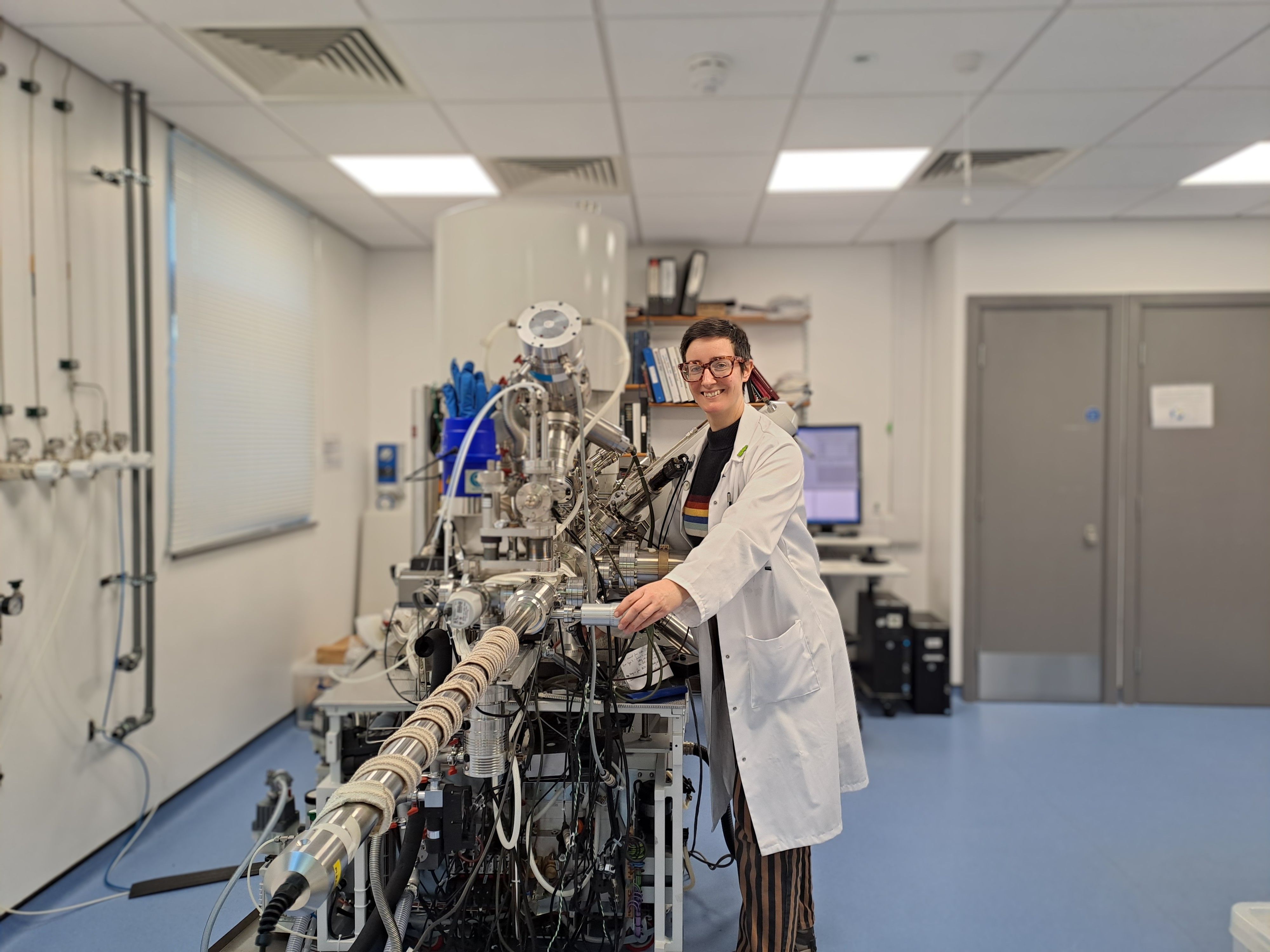 Dr Hannah Constantin, a Materials Characterisation Research Technician at University of Nottingham, stands beside a large materials analysis instrument in a laboratory, smiling as she adjusts part of the complex equipment. Wearing a lab coat and surrounded by cables, monitors, and vacuum chambers, she works with advanced surface analysis technology used to study the properties and composition of materials.