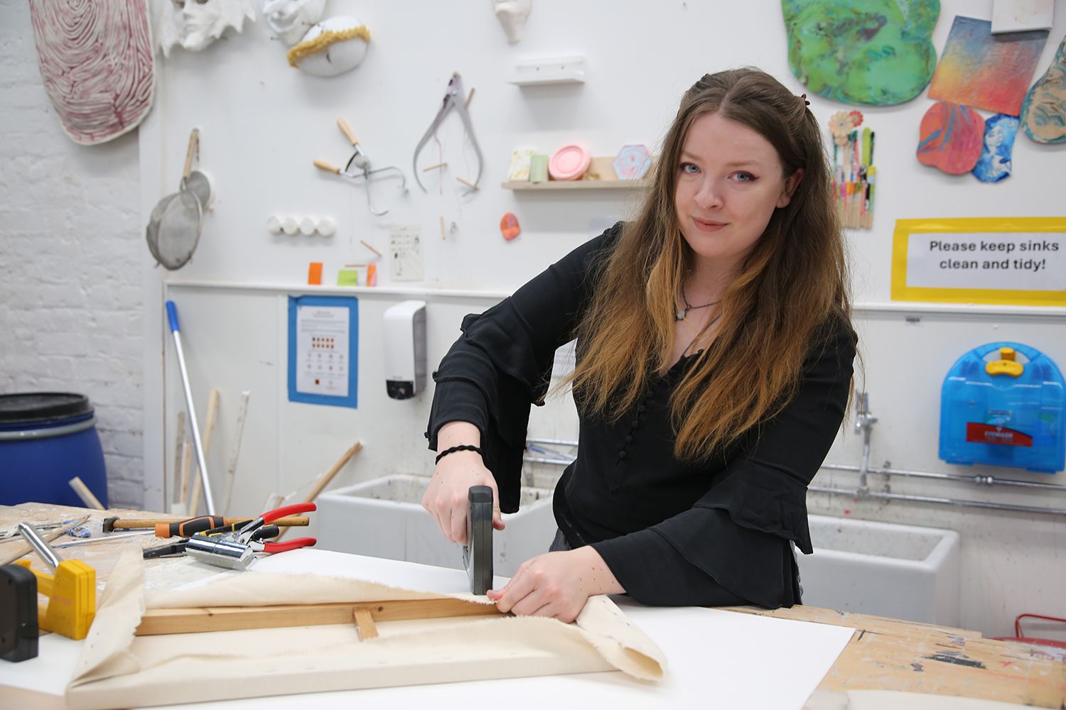 Meg Golding, Technical Demonstrator in the Institute of Arts and Humanities at the University of Worcester, works at a studio workbench, stretching and securing canvas over a wooden frame using a staple gun. Surrounded by tools, materials, and colourful artworks on the walls, she demonstrates practical techniques that support students in developing their creative practice.