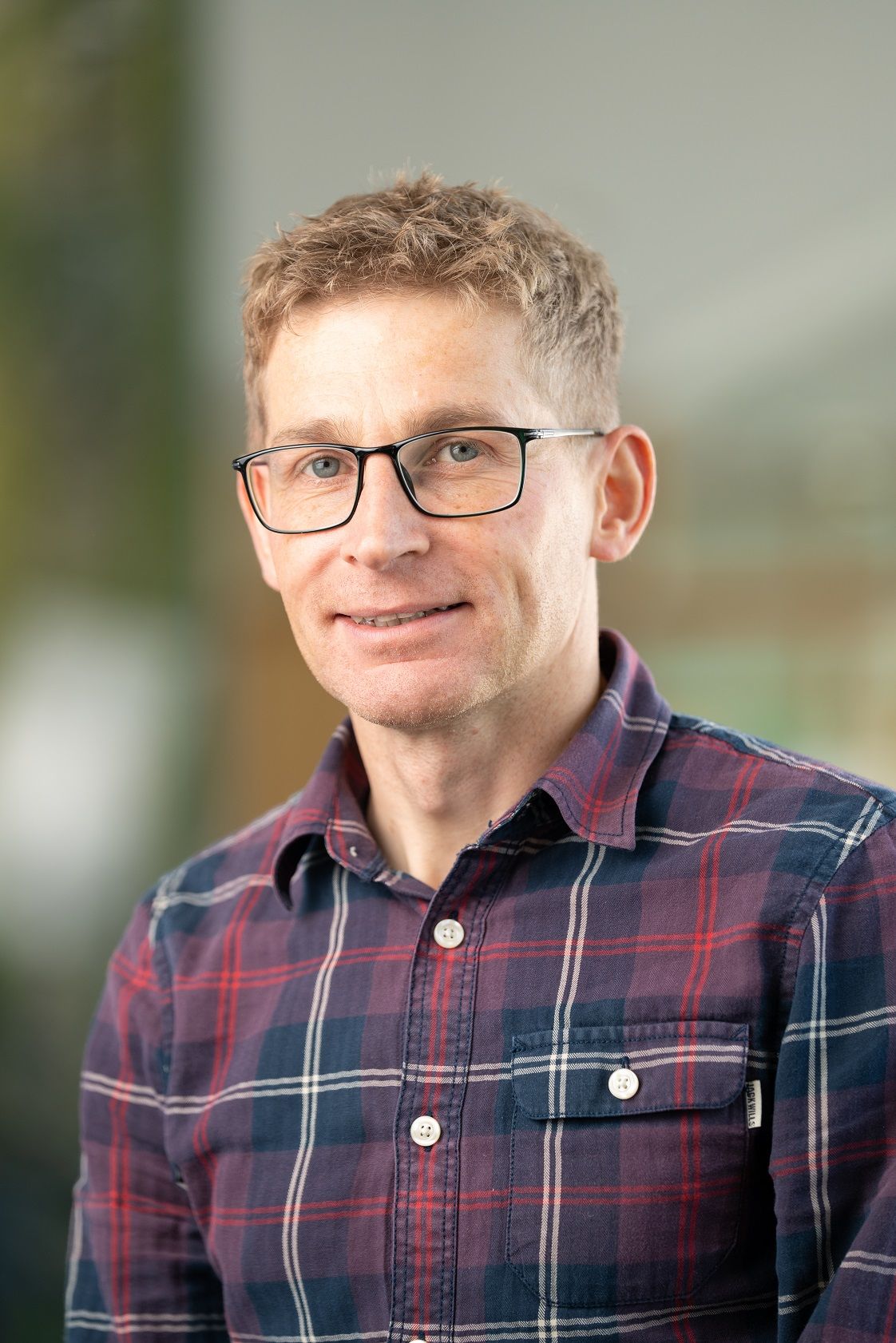 Doctor Alex Laude, Head of Biomedical Research Facilities at Newcastle University, smiles in a professional portrait setting. He is wearing glasses and a red and navy checked shirt. Dr Laude is one of eight nominees shortlisted for the 2025 Times Higher Education Outstanding Technician of the Year award.