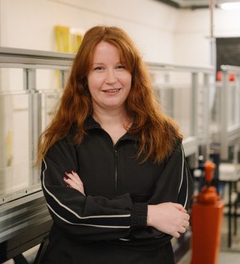 Helena Brown, Technical Specialist in the School of Earth and Environment at the University of Leeds, stands with her arms crossed in a laboratory setting. She has long red hair and is wearing a black zip-up top with white stripes on the sleeves. Helena is one of eight nominees shortlisted for the 2025 Times Higher Education Outstanding Technician of the Year award.