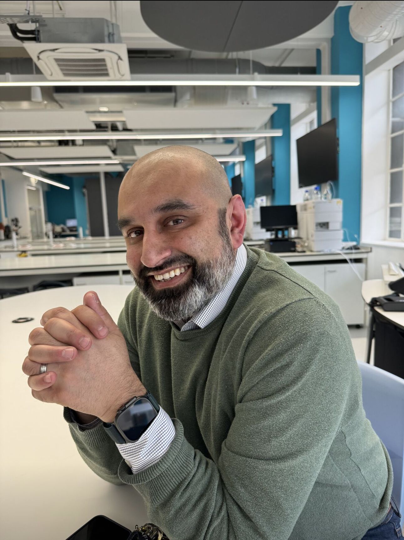 Jiteen Ahmed, Head of Technical Services in the College of Health and Life Sciences at Aston University, smiles while seated at a table in a bright laboratory setting. He is wearing a green jumper over a collared shirt and an Apple Watch. Jiteen has a neatly trimmed beard and is one of eight nominees shortlisted for the 2025 Times Higher Education Outstanding Technician of the Year award.