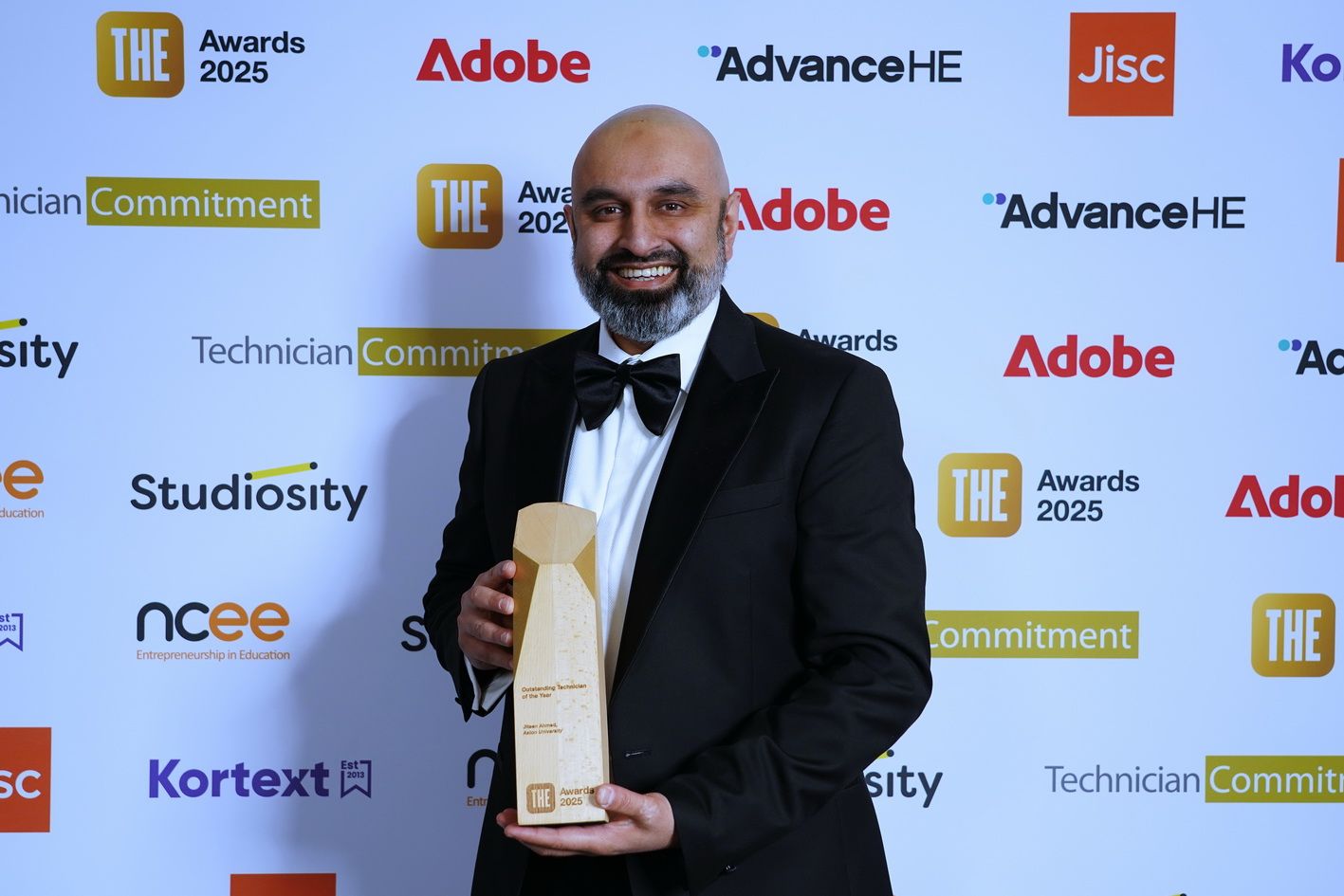 Jiteen Ahmed, Head of Technical Services in the College of Health and Life Sciences at Aston University, smiles while holding his trophy after being named the winner of the Times Higher Education Outstanding Technician of the Year Award. He is wearing a black tuxedo and bow tie, standing in front of a branded backdrop featuring logos from sponsors including Adobe, Advance HE, and Technician Commitment, at the celebration event held at Edinburgh Conference Centre on Thursday 13 November 2025.
