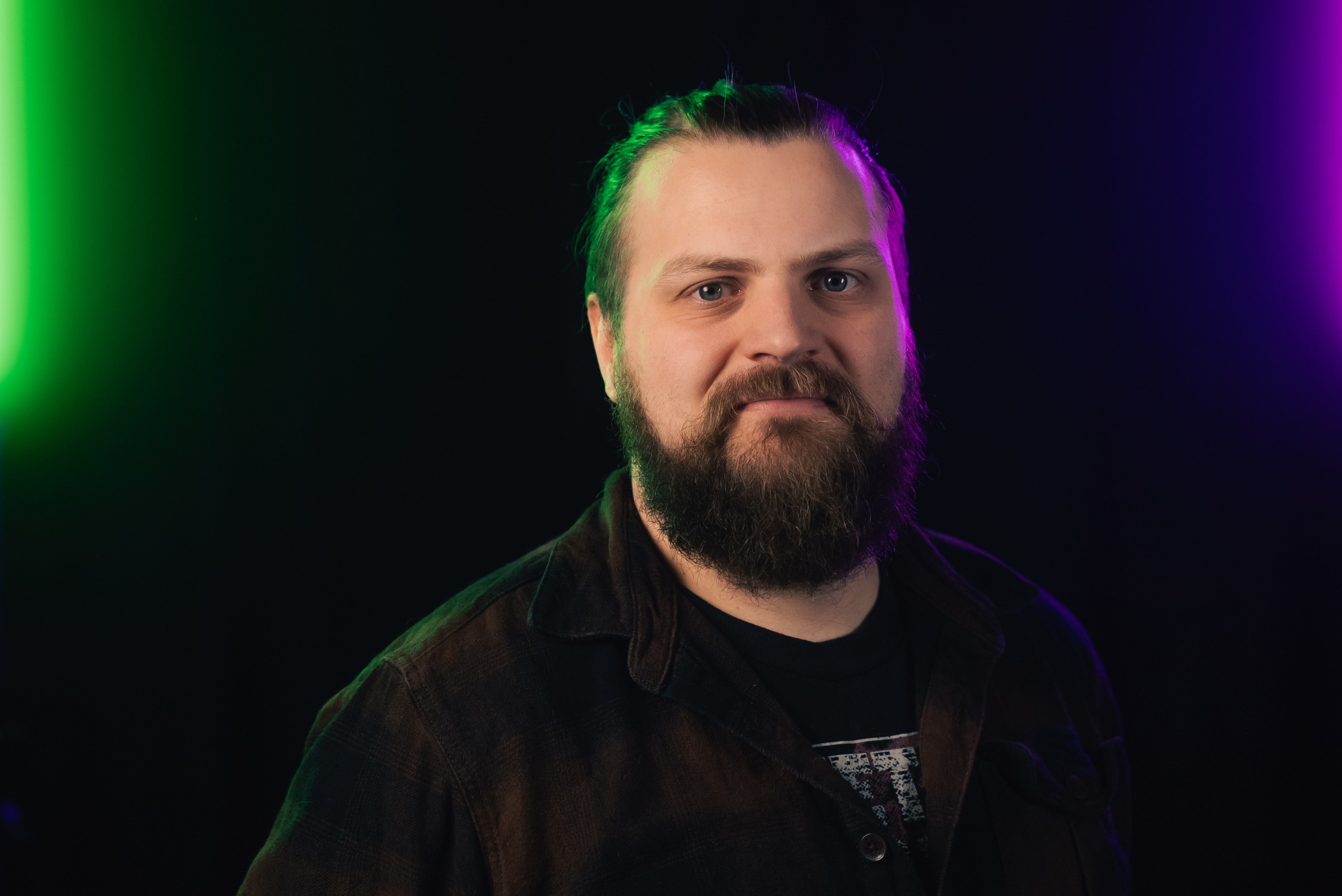 Joe Rees-Jones, Research Technician in the School of Physics, Engineering and Technology at the University of York, poses against a dark background illuminated by green and purple lighting. He has a beard and his hair tied back, wearing a dark shirt and T-shirt. Joe is one of eight nominees shortlisted for the 2025 Times Higher Education Outstanding Technician of the Year award.
