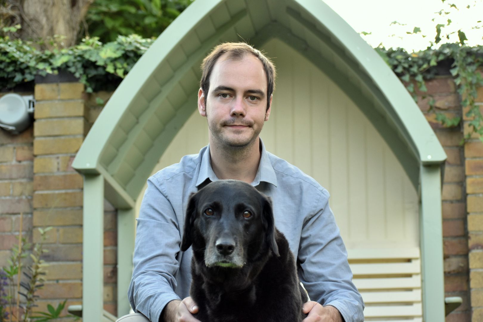 John Nicolson, Technician Development Adviser at the University of Cambridge, sits outdoors on a garden bench with a black Labrador resting on his lap. He is wearing a light blue shirt and looking toward the camera. John is one of eight nominees shortlisted for the 2025 Times Higher Education Outstanding Technician of the Year award.