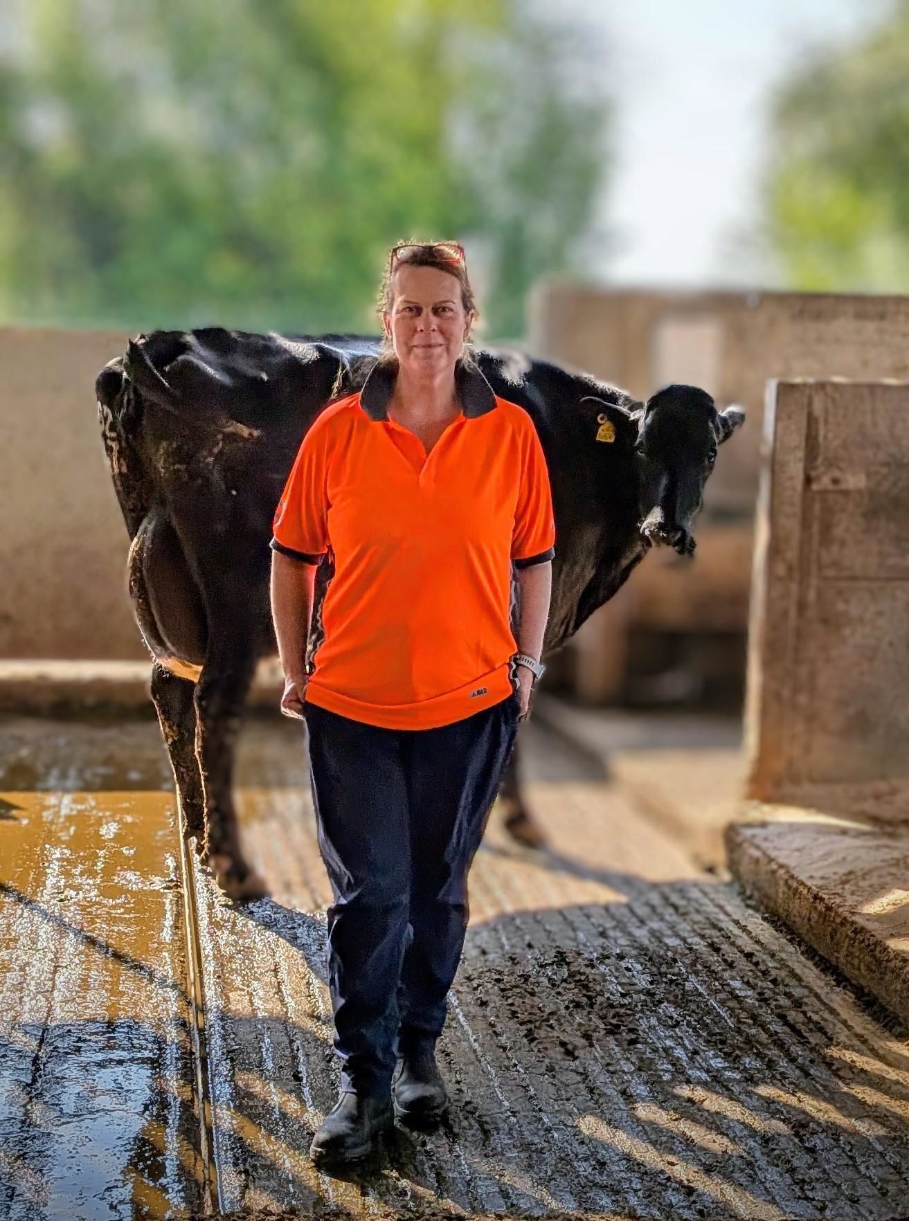 Kate Robinson, Ruminant Section Manager at Harper Adams University’s Future Farm, stands in a barn with a black cow behind her. She is wearing a bright orange polo shirt and dark trousers, smiling slightly while posing on the farm walkway. Kate is one of eight nominees shortlisted for the 2025 Times Higher Education Outstanding Technician of the Year award.