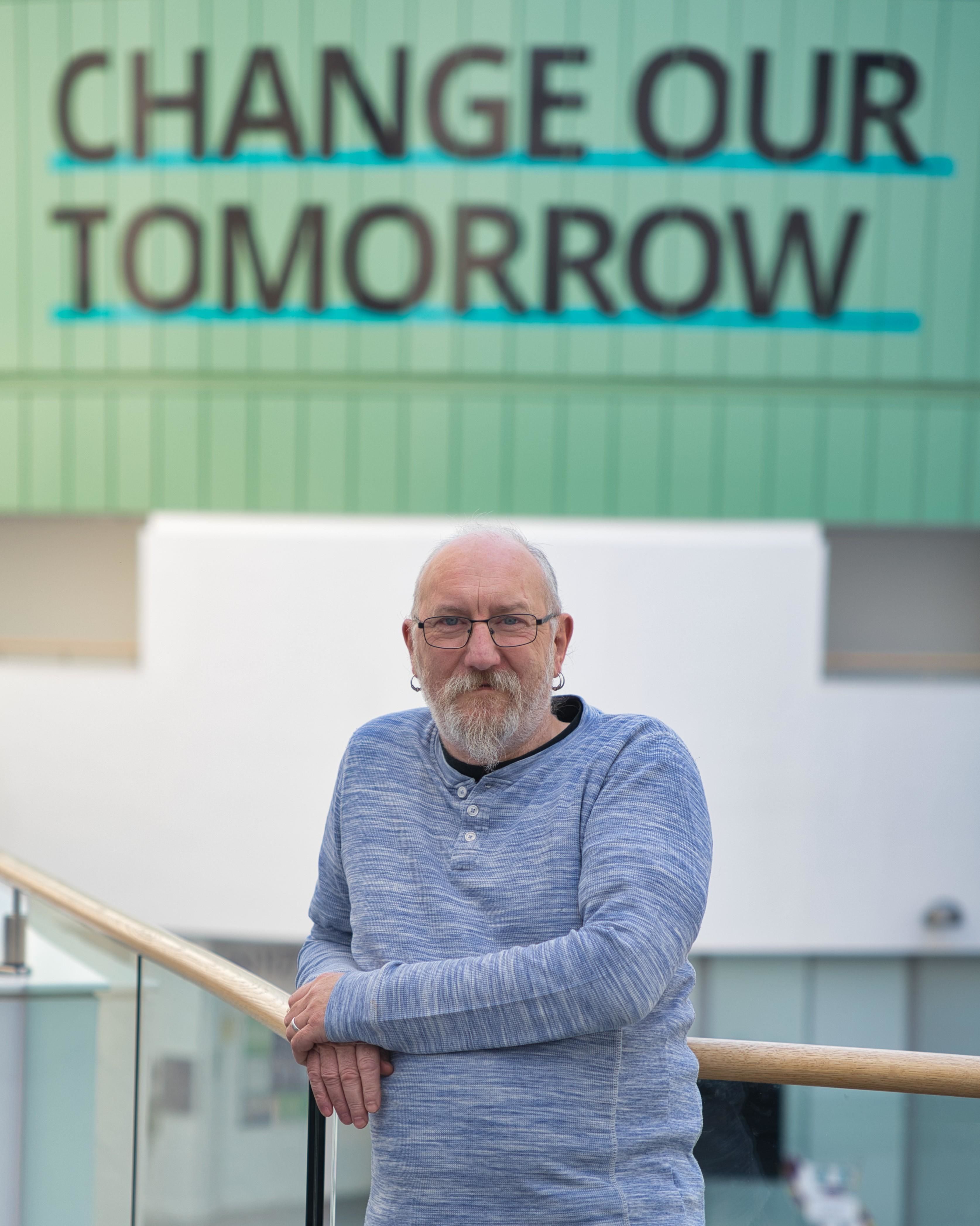 Phil Chaplain, Engineering Services Manager and Health & Safety Coordinator in the School of Computing, Engineering & Technology at Robert Gordon University, stands leaning on a glass railing inside a modern university building. He is wearing a light blue long-sleeved top and glasses, with the words “Change Our Tomorrow” visible on the wall behind him. Phil is one of eight nominees shortlisted for the 2025 Times Higher Education Outstanding Technician of the Year award.
