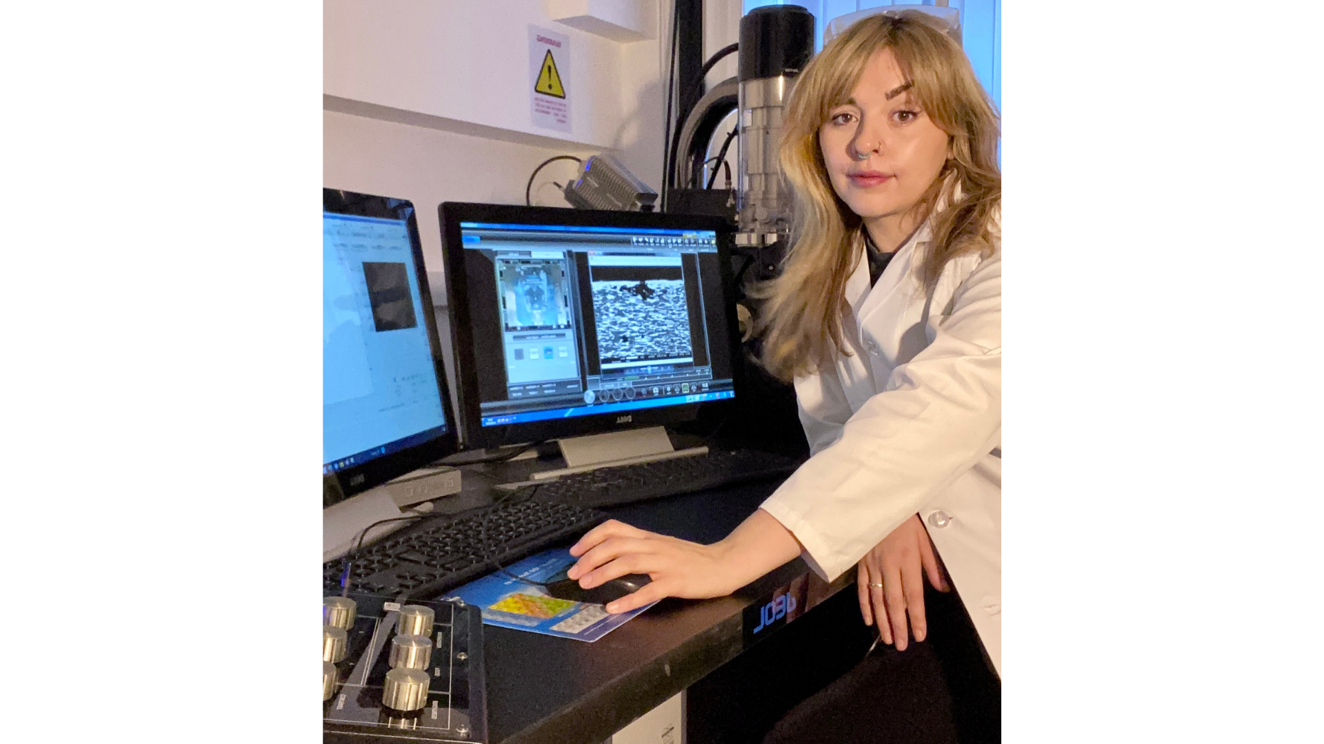 Dr Nicola George, Research Technical Professional for Archaeological Science and Facility Manager, Professor Elizabeth Slater Archaeological Research Laboratories, University of Liverpool, stands at a workstation beside a high-powered microscope and analysis system, reviewing detailed imaging data on dual computer monitors. In the laboratory, she supports archaeological science research by managing specialist equipment and analysing microscopic evidence from archaeological materials.