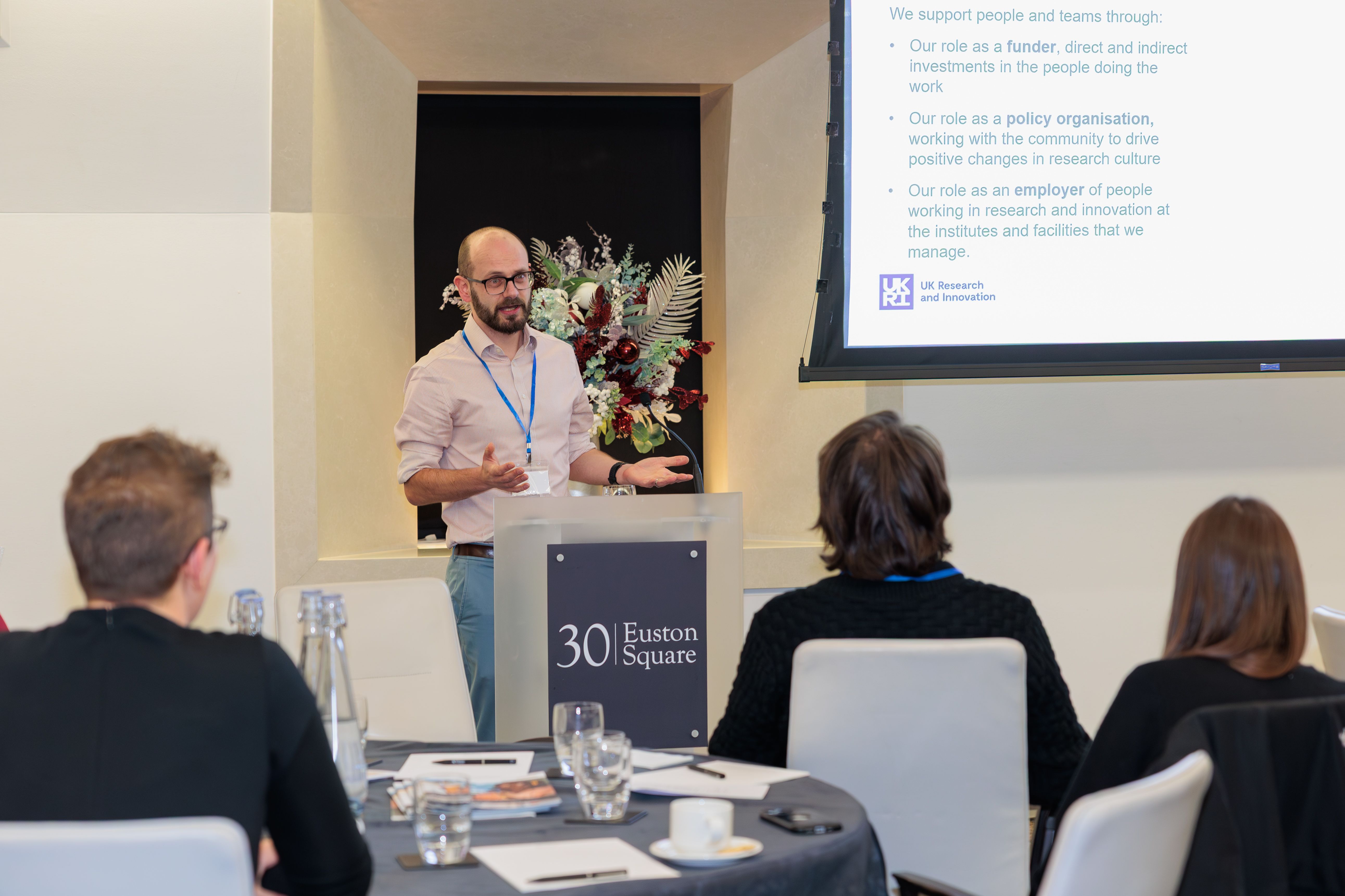 Nik Ogryzko, Senior Talent Programme Manager at UKRI standing at a podium speaking into a microphone in front of a PowerPoint slide that reads 'We support people and teams through: • Our role as a funder, direct and indirect investments in the people doing the work • Our role as a policy organisation, working with the community to drive positive changes in research culture • Our role as an employer of people working in research and innovation at the institutes and facilities that we manage.' The UKRI logo is visible in the bottom left-hand corner of the image. The heads of audience members can be seen in the foreground turned towards Nik as he speaks.