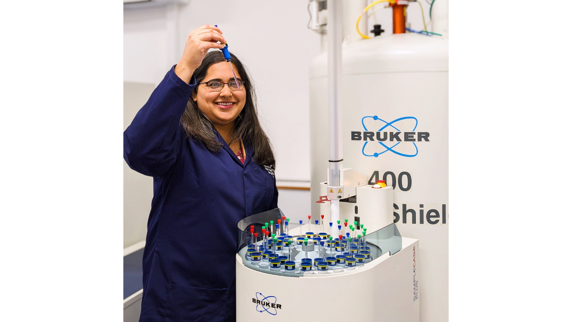 Nikita Harvey, School of Pharmacy NMR Facility Manager, UCL, stands beside an NMR spectrometer in a laboratory, smiling as she carefully holds a small sample tube above a rack of prepared samples. Wearing a lab coat and glasses, she works with precision equipment used to analyse the chemical structure of compounds, highlighting the technical expertise required to support advanced research in pharmaceutical science.