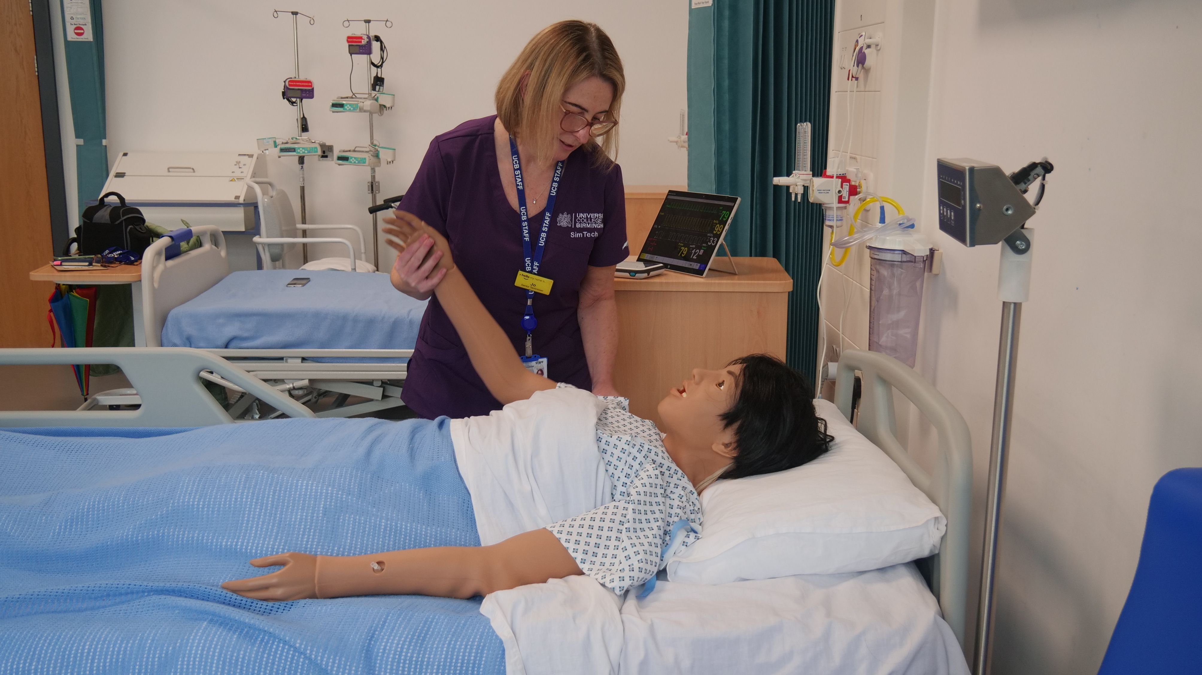 Jo Simmons, Senior Technician – Clinical Skills and Simulation in the School of Health, Life Sciences and Education, University College Birmingham stands beside a hospital bed in a clinical training suite, demonstrating patient care techniques by gently raising the arm of a life-sized medical simulation mannequin. Wearing purple clinical scrubs and a staff lanyard, she focuses attentively on the task, surrounded by hospital equipment used to train healthcare students in realistic clinical scenarios.