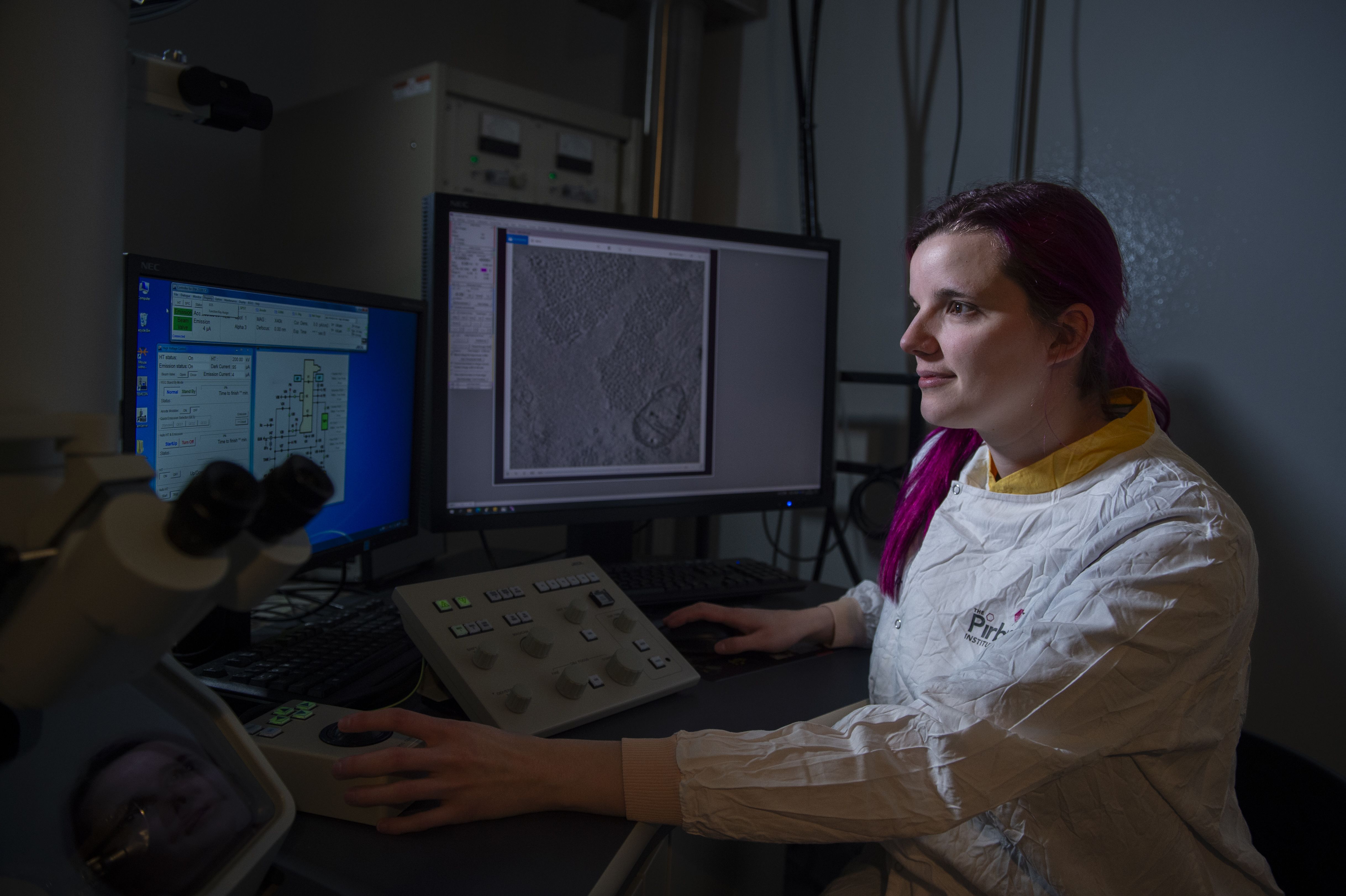 Joanna Wells, Microscopist at The Pirbright Institute, sits at a specialised microscope workstation, carefully controlling imaging equipment while examining a sample displayed on nearby computer screens. Wearing a protective laboratory suit, she focuses on high-resolution microscopy used to study biological samples and support research into infectious animal diseases.