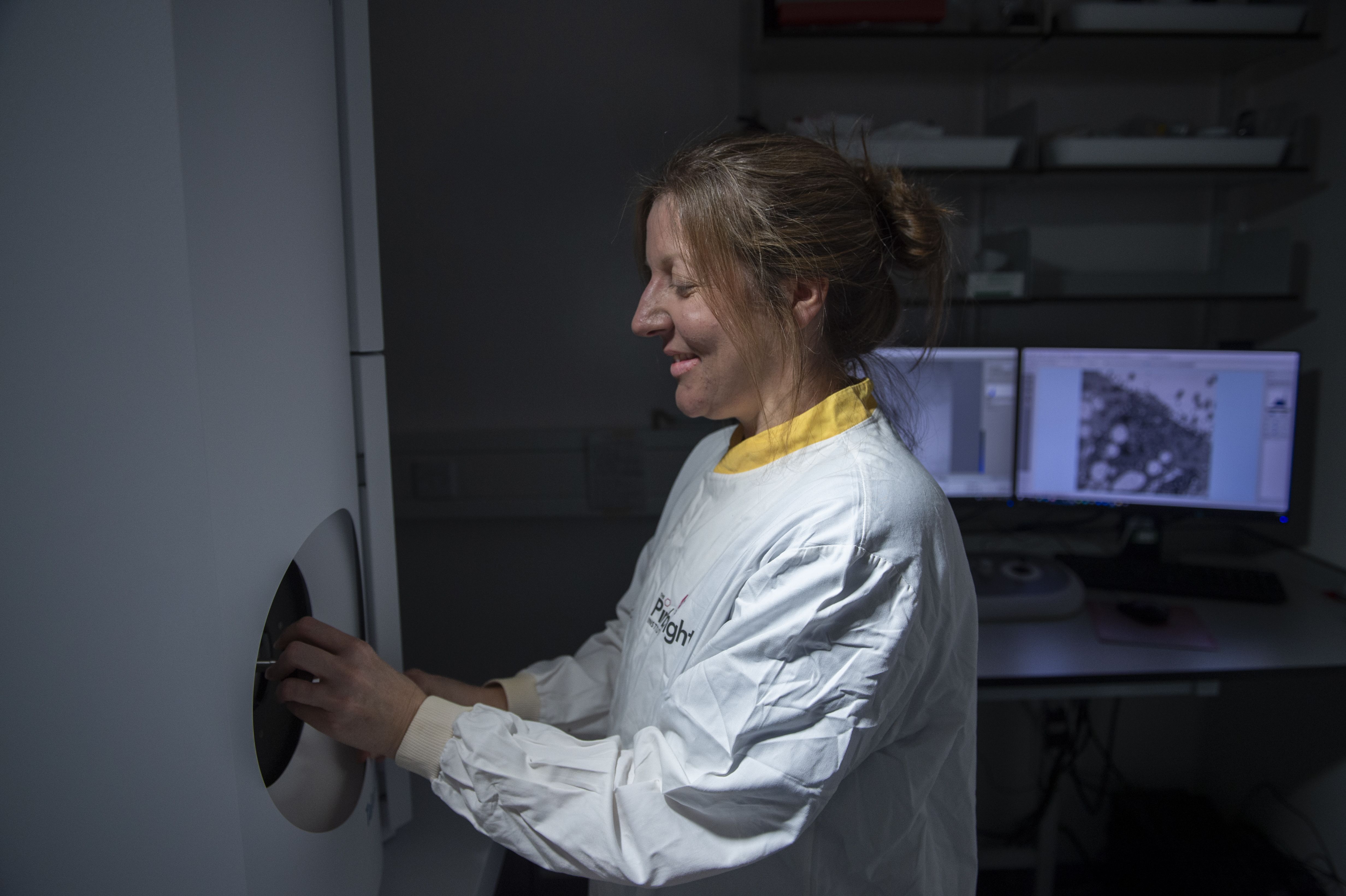 Jennifer Simpson, Head of Bioimaging at The Pirbright Institute, stands beside a high-resolution imaging instrument, carefully placing a sample into the machine while wearing a protective laboratory suit. Behind her, computer monitors display detailed microscope images used to analyse biological samples and support research into infectious diseases.