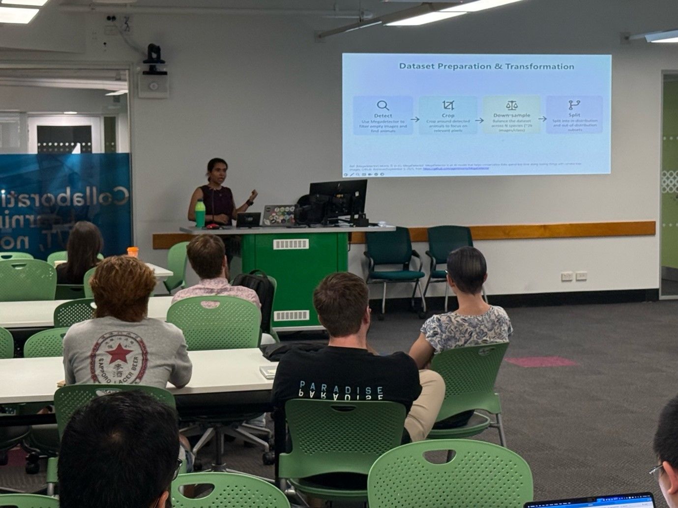 Dr Renuka Sharma, Data Scientist at QCIF Digital Research, stands at the front of a lecture room delivering a presentation, gesturing toward a projected slide that explains steps in data preparation and transformation. Speaking to an audience seated at tables, she shares insights from her work in data science and digital research.