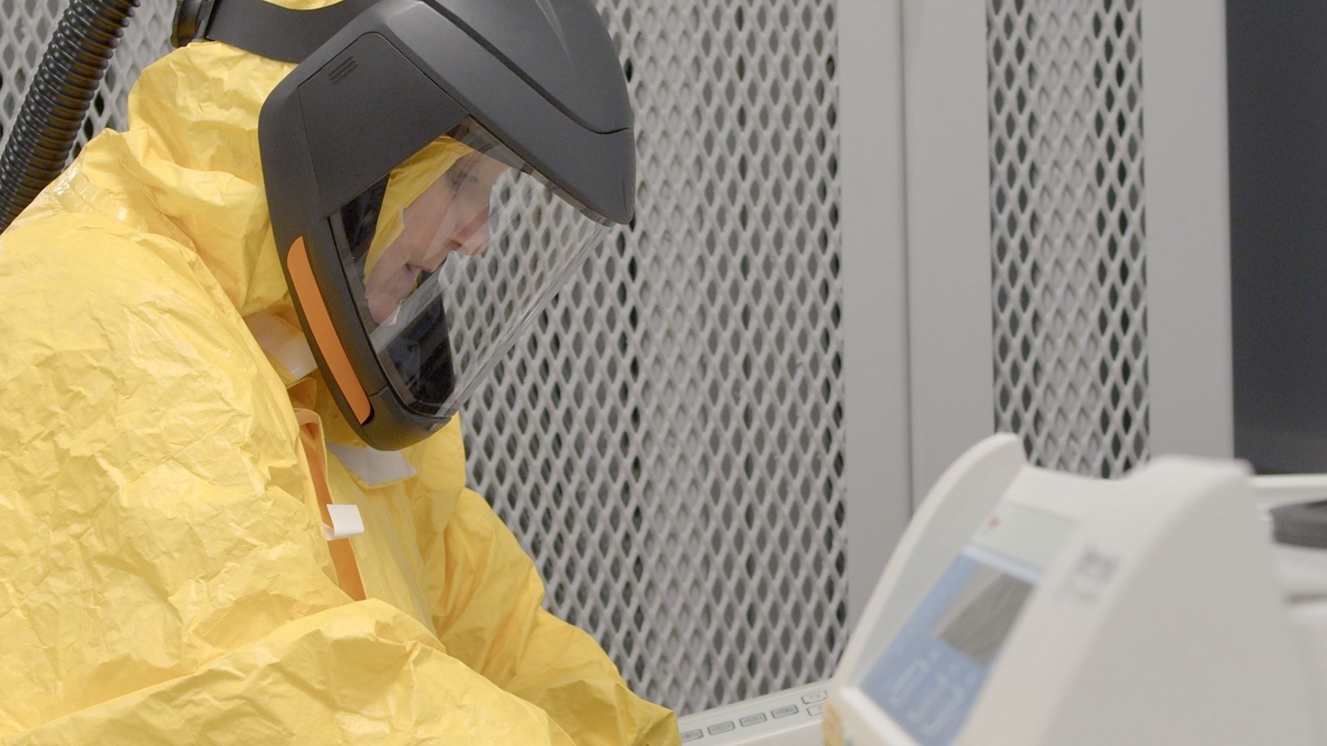 Julia Spoors, Infectious Diseases Facility (CL3) Manager, Newcastle University works inside a high-containment laboratory wearing full protective equipment, including a sealed respirator helmet and a yellow biosafety suit. Focused on the task in front of her, she operates specialised laboratory equipment designed for safely handling infectious disease samples in a controlled environment.