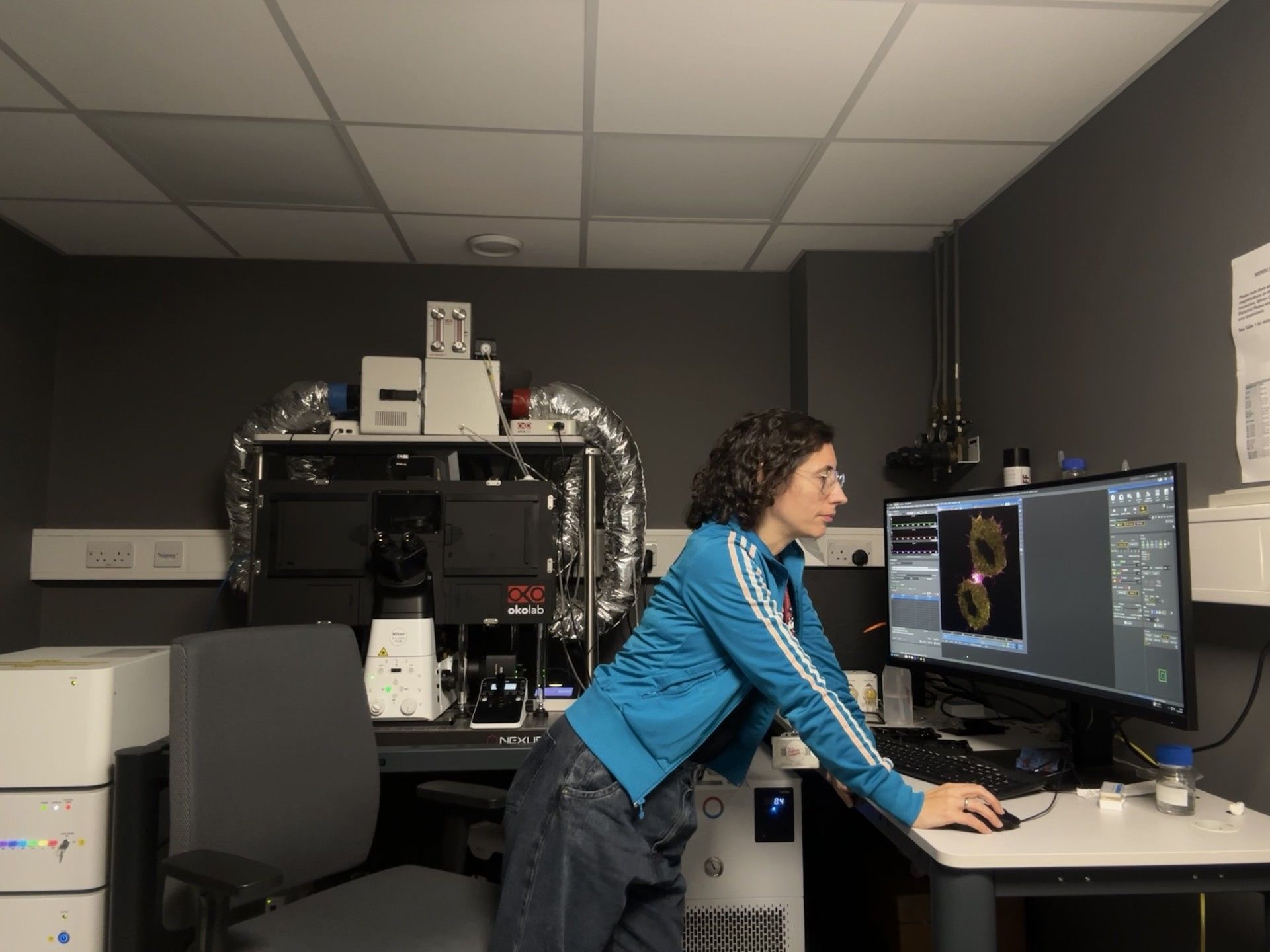 Virginia Silió, Imaging Technical Specialist at University College London, stands at a computer workstation beside a high-resolution microscope system, studying detailed cellular images displayed on the monitor. Surrounded by advanced imaging equipment in a specialised microscopy suite, she analyses biological samples to support cutting-edge research.